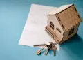 A small wooden model house sits on a document with text about ways to save on home insurance, accompanied by a set of brass keys on a keyring. The scene is set against a blue background. | MyWebInsurance