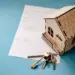 A small wooden model house sits on a document with text about ways to save on home insurance, accompanied by a set of brass keys on a keyring. The scene is set against a blue background. | MyWebInsurance