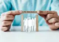 Two hands hold up wooden blocks with the words "Life Insurance," forming a frame over white silhouettes of multiple people standing close together, illustrating the concept of life insurance coverage—be it Term vs Whole Life Insurance—protecting a group of individuals. | MyWebInsurance