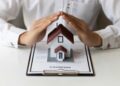 A person places their hands protectively over a small model house situated on a document labeled "Renters Insurance." The document is held down with a clipboard on a white surface. The individual is dressed in a white shirt, emphasizing the concept of home and renters insurance. | MyWebInsurance