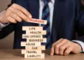 A person in a suit and tie is playing a game of Jenga with wooden blocks labeled with various types of insurances like life, health, legal expenses, business, house, car, travel, and liability. The person is placing a block labeled "INSURANCE" on top of the stack. | MyWebInsurance