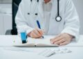 A person in a white lab coat and stethoscope sits at a desk in a medical office, writing in a notebook with a blue pen, perhaps noting tips on how to choose health insurance. Nearby are a blue stamp, a blister pack of pills, and a syringe. | MyWebInsurance