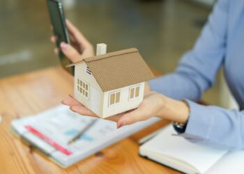 A person in business attire holds a small model of a house in one hand and a smartphone in the other. On the wooden table in front of them are documents and an open notebook, highlighting concepts of real estate or home insurance. | MyWebInsurance