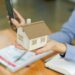 A person in business attire holds a small model of a house in one hand and a smartphone in the other. On the wooden table in front of them are documents and an open notebook, highlighting concepts of real estate or home insurance. | MyWebInsurance