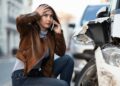 A worried woman crouches beside a damaged car, holding her head with one hand and speaking on her phone with the other. She wears a brown leather jacket and jeans, and the car's front is severely dented. As she assesses the damage, she seems to wonder, "How much auto insurance do I need?" A street and other vehicles are visible in the background. | MyWebInsurance