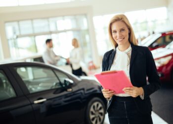 A woman with blonde hair, dressed in business attire, stands smiling while holding a pink folder in a car showroom. In the background, a man and woman are engaged in conversation about Car Insurance Florida near other vehicles. The showroom is well-lit and bright. | MyWebInsurance