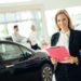 A woman with blonde hair, dressed in business attire, stands smiling while holding a pink folder in a car showroom. In the background, a man and woman are engaged in conversation about Car Insurance Florida near other vehicles. The showroom is well-lit and bright. | MyWebInsurance