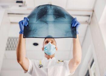 A healthcare professional wearing a mask and blue gloves holds an X-ray image of a chest up to the light. The X-ray shows the ribcage and lungs. The individual, covered by extensive health insurance, is wearing a white coat and standing in a bright room. | MyWebInsurance