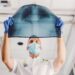 A healthcare professional wearing a mask and blue gloves holds an X-ray image of a chest up to the light. The X-ray shows the ribcage and lungs. The individual, covered by extensive health insurance, is wearing a white coat and standing in a bright room. | MyWebInsurance