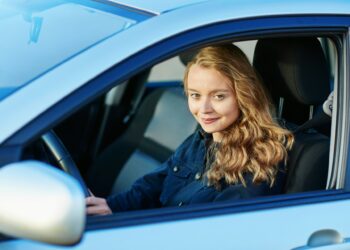 A young woman with long, wavy blond hair is sitting in the driver's seat of a blue car, looking at the camera and smiling. She is wearing a dark jacket, her hands on the steering wheel, embodying the confidence often seen with those who benefit from good Car Insurance Young Drivers. | MyWebInsurance