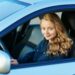 A young woman with long, wavy blond hair is sitting in the driver's seat of a blue car, looking at the camera and smiling. She is wearing a dark jacket, her hands on the steering wheel, embodying the confidence often seen with those who benefit from good Car Insurance Young Drivers. | MyWebInsurance