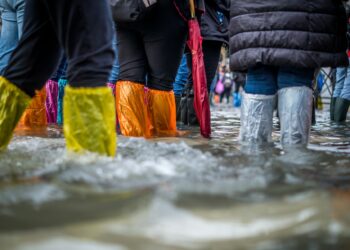 People walking through a flood wearing colorful plastic boot covers, including yellow, orange, and silver. The water is ankle-deep, reflecting the boots and clothing of the individuals as they navigate the flooded area caused by water damage. | MyWebInsurance