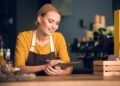 A woman in a yellow shirt and brown apron smiles while holding a clipboard and pen, perhaps checking off items on a business insurance checklist. She stands behind a wooden counter, with bottles and cups on shelves in the background, suggesting a cozy cafe or restaurant setting. | MyWebInsurance