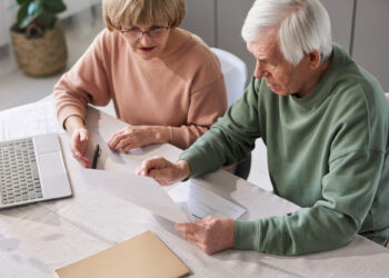 An elderly couple sits at a table, reviewing Affordable Care Act documents. The woman wears glasses and a pink sweater, while the man dons green. A laptop and folder are on the table, with a plant visible in the background. | MyWebInsurance