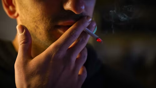 A close-up of a person holding a lit cigarette near their mouth, with smoke visible and the background blurred. The image captures the details of their hand and cigarette, subtly highlighting smoking habits. | MyWebInsurance