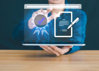 A person in a blue shirt holds a laptop displaying holographic icons of a certificate with a check mark and a signed document, symbolizing digital certification or online document verification. | MyWebInsurance