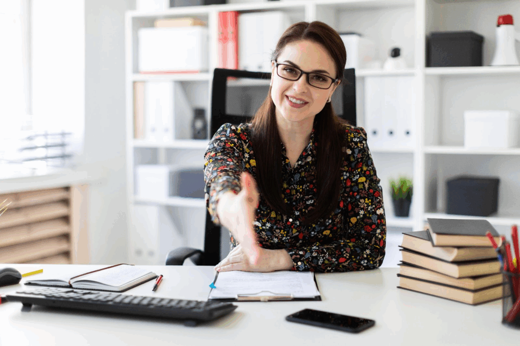 A woman with glasses, wearing a floral blouse, sits at a desk, smiling and reaching out for a handshake. Books, a notepad, a pen, and a smartphone are on the desk, with shelves in the background. | MyWebInsurance