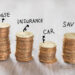 Four stacks of coins labeled "House," "Insurance," "Car," and "Savings" on a wooden surface, illustrating different financial categories or budgeting goals. | MyWebInsurance