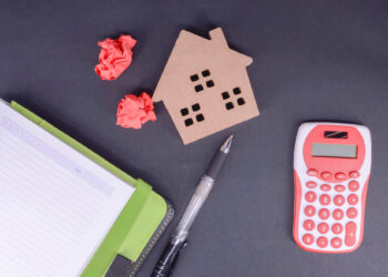 Flat lay of a notepad, pen, two red crumpled paper balls, a small wooden house cutout, and a red-and-white calculator on a dark background. | MyWebInsurance