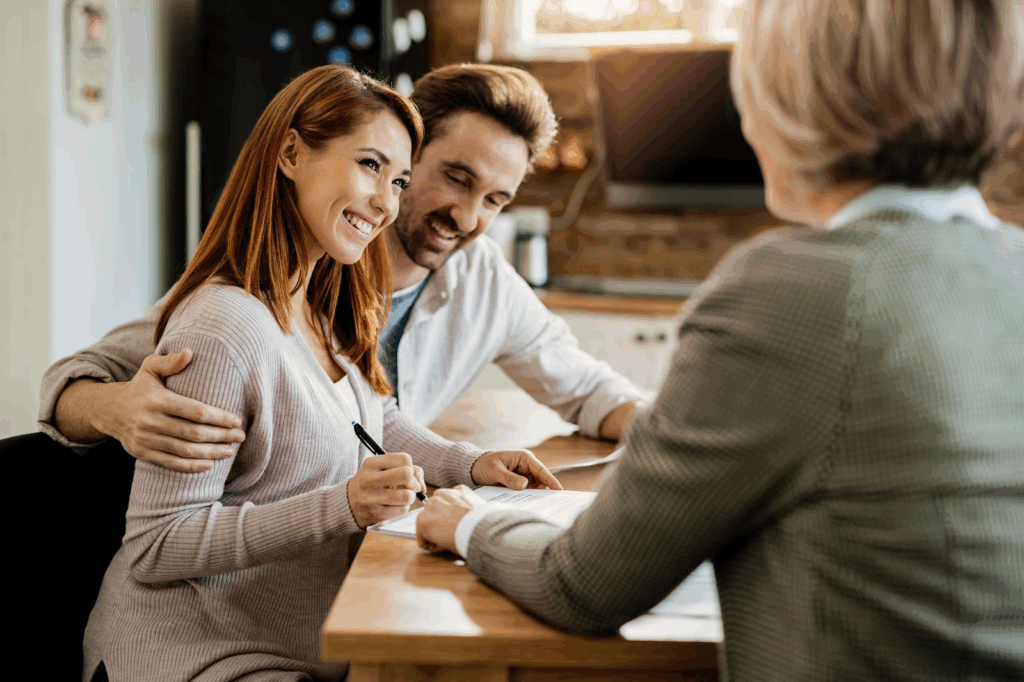 A smiling couple sits at a table with a woman, signing documents. The man has his arm around the woman, and they appear happy and engaged during the meeting in a bright, homey setting. | MyWebInsurance