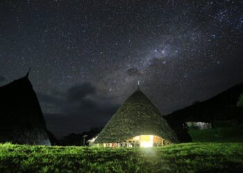 A thatched hut glows with warm light beneath a star-filled night sky, surrounded by grass and distant hills. The Milky Way is clearly visible overhead. | MyWebInsurance