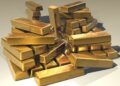A large pile of shiny gold bars stacked haphazardly on a neutral surface, reflecting light and showing some surface scratches. | MyWebInsurance