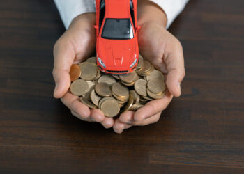 A person holds a pile of coins in their cupped hands, with a small red toy car placed on top, all set against a dark wooden surface. | MyWebInsurance