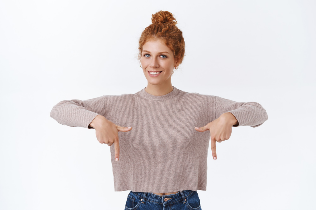 A woman with curly red hair in a bun, wearing a beige long-sleeve shirt and jeans, smiles while pointing down with both hands against a plain white background. | MyWebInsurance