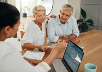 An older couple sits at a table, attentively listening to a professional who is showing them information on a laptop and holding documents, suggesting a financial or retirement planning discussion. | MyWebInsurance
