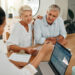 An older couple sits at a table, attentively listening to a professional who is showing them information on a laptop and holding documents, suggesting a financial or retirement planning discussion. | MyWebInsurance