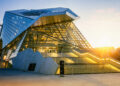 A modern building with angular glass and metal architecture, wide illuminated stairs leading to the entrance, and sunlight shining in the background. | MyWebInsurance
