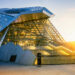 A modern building with angular glass and metal architecture, wide illuminated stairs leading to the entrance, and sunlight shining in the background. | MyWebInsurance
